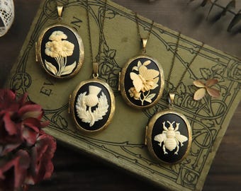 Puede incluir: Cuatro relicarios ovalados de estilo antiguo con diseños florales e insectos. Cada relicario presenta un fondo negro con un diseño tallado en color crema de una flor, una mariposa, un cardo y una abeja. Los relicarios cuelgan de cadenas de color bronce.