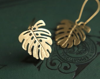 May include: A pair of gold-tone earrings shaped like monstera leaves. The earrings are delicate and have a brushed finish.