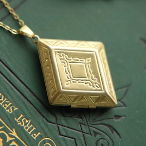 May include: A gold locket necklace with a diamond-shaped pendant featuring an intricate engraved design. The locket is closed and has a hinge on the side.