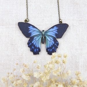 K&ouml;nnte beinhalten: Eine Halskette mit Schmetterlingsanh&auml;nger und einem dunkelblauen und hellblauen Schmetterlingsdesign. Der Schmetterlingsanh&auml;nger h&auml;ngt an einer filigranen Kette. Die Halskette wird vor einem wei&szlig;en, strukturierten Hintergrund mit kleinen wei&szlig;en Blumen pr&auml;sentiert.