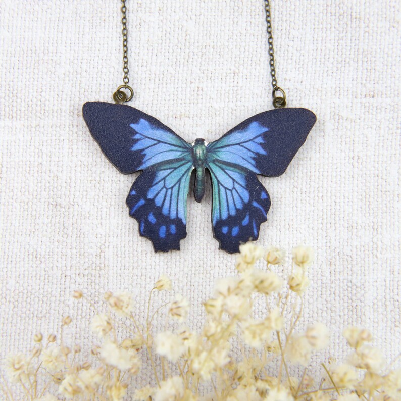 K&ouml;nnte beinhalten: Eine Halskette mit Schmetterlingsanh&auml;nger und einem dunkelblauen und hellblauen Schmetterlingsdesign. Der Schmetterlingsanh&auml;nger h&auml;ngt an einer filigranen Kette. Die Halskette wird vor einem wei&szlig;en, strukturierten Hintergrund mit kleinen wei&szlig;en Blumen pr&auml;sentiert.