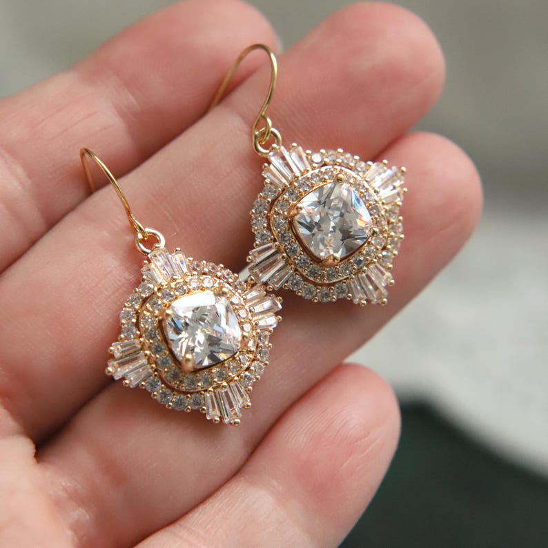 May include: A pair of gold-toned drop earrings. Each earring features a square, clear gemstone at the center, surrounded by smaller, clear gemstones in a halo and baguette arrangement. The earrings have a classic, elegant design.