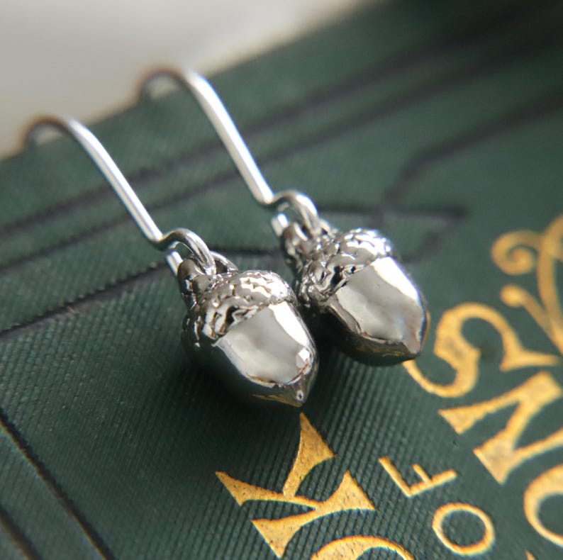 May include: A pair of silver-toned metal earrings shaped like small acorns. Each earring features a textured cap and a smooth, polished nut body, suspended from a simple curved silver-toned ear wire hook for attachment to the earlobe.