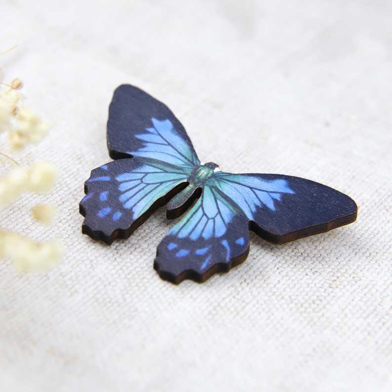 K&ouml;nnte beinhalten: Ein Holzschmetterling mit detailliertem Design. Der Schmetterling hat einen dunkelblauen K&ouml;rper und Fl&uuml;gel mit blau-t&uuml;rkisfarbenen Mustern. Die Fl&uuml;gel sind aufwendig geformt, und der Schmetterling ist vor einem neutralen Hintergrund platziert.