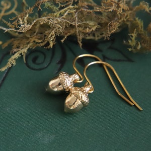 May include: A pair of gold-toned drop earrings featuring small, detailed acorn charms. Each acorn has a smooth, polished nut section and a textured, cap-like top. The charms are suspended from simple, curved gold-toned ear wires for a classic look.