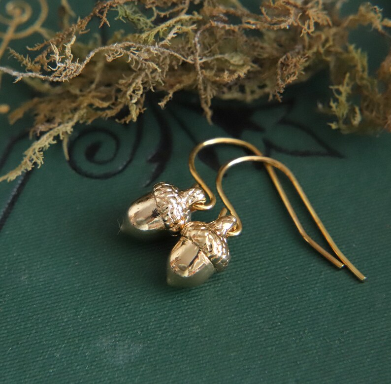 May include: A pair of gold-toned drop earrings featuring small, detailed acorn charms. Each acorn has a smooth, polished nut section and a textured, cap-like top. The charms are suspended from simple, curved gold-toned ear wires for a classic look.
