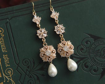 May include: A pair of gold-toned drop earrings featuring a linear design with clear baguette-cut crystals, a central floral-shaped component encrusted with small clear stones, and a single white teardrop-shaped faux pearl at the base of each earring.