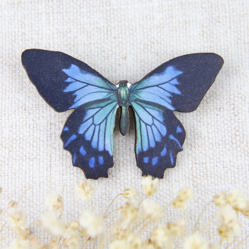 K&ouml;nnte beinhalten: Ein Holzschmetterling mit einem detaillierten blau-schwarzen Fl&uuml;gelmuster. Der Schmetterling hat einen dunkelblauen K&ouml;rper und Fl&uuml;gel mit verschiedenen Blaut&ouml;nen. Die Fl&uuml;gel sind ausgebreitet und zeigen das komplizierte Design.