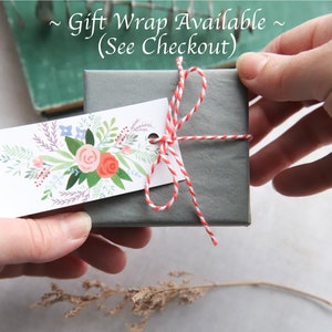Puede incluir: Una caja de regalo gris atada con cordel de panadero rojo y blanco y una etiqueta de regalo floral. La etiqueta dice "Gift Wrap Available (See Checkout)".