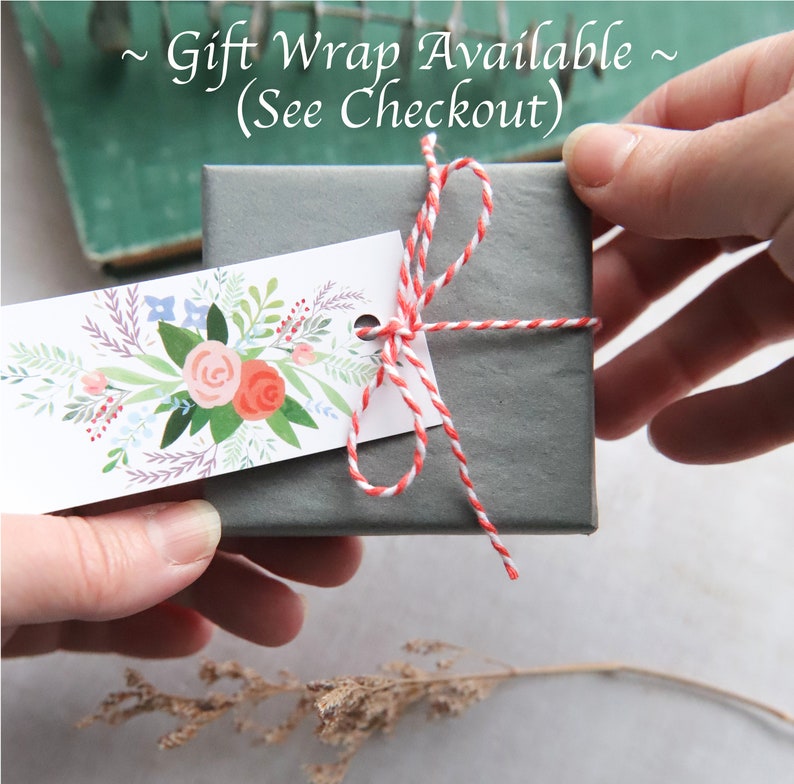 Puede incluir: Una caja de regalo gris atada con cordel de panadero rojo y blanco y una etiqueta de regalo floral. La etiqueta dice "Gift Wrap Available (See Checkout)".