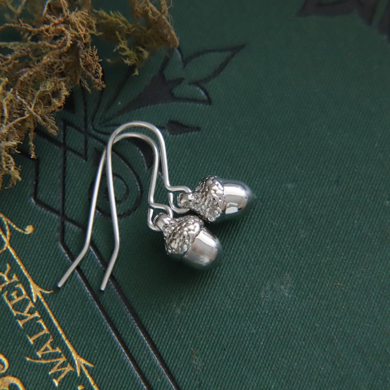 May include: A pair of silver-toned acorn earrings featuring textured caps and smooth, polished nut bodies. The earrings are attached to simple silver-toned ear wires, showcasing a detailed, realistic design that captures the natural shape of an acorn.