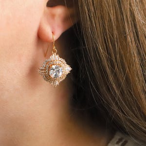 May include: A gold-toned earring with a square, clear center stone and a surrounding design of smaller, clear stones. The earring has a decorative, geometric shape and a hook closure. The earring is shown against a blurred background.