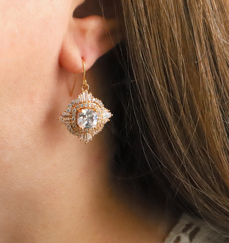 May include: A gold-toned earring with a square, clear center stone and a surrounding design of smaller, clear stones. The earring has a decorative, geometric shape and a hook closure. The earring is shown against a blurred background.
