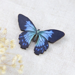 K&ouml;nnte beinhalten: Ein Holzschmetterling mit detaillierten Fl&uuml;geln in Blau- und Schwarzt&ouml;nen. Der Schmetterling, m&ouml;glicherweise eine Brosche oder Verzierung, ist vor einem neutralen Hintergrund platziert.