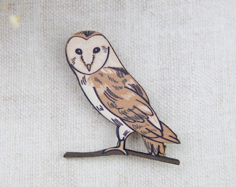 Puede incluir: Un broche de madera con forma de búho de granero con detalles pintados en marrón y blanco. El búho está posado en una rama con las alas extendidas.