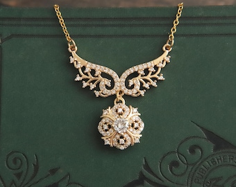 May include: A gold-toned necklace with a floral pendant and a delicate chain. The pendant features a central clear stone surrounded by smaller stones, with an ornate design. The necklace has a decorative, wing-like upper section, and is set against a dark green background.