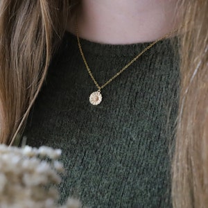 Puede incluir: Un collar de cadena dorada con un delicado colgante de margarita. El collar lo lleva una persona con el pelo largo y casta&ntilde;o y un jersey verde.