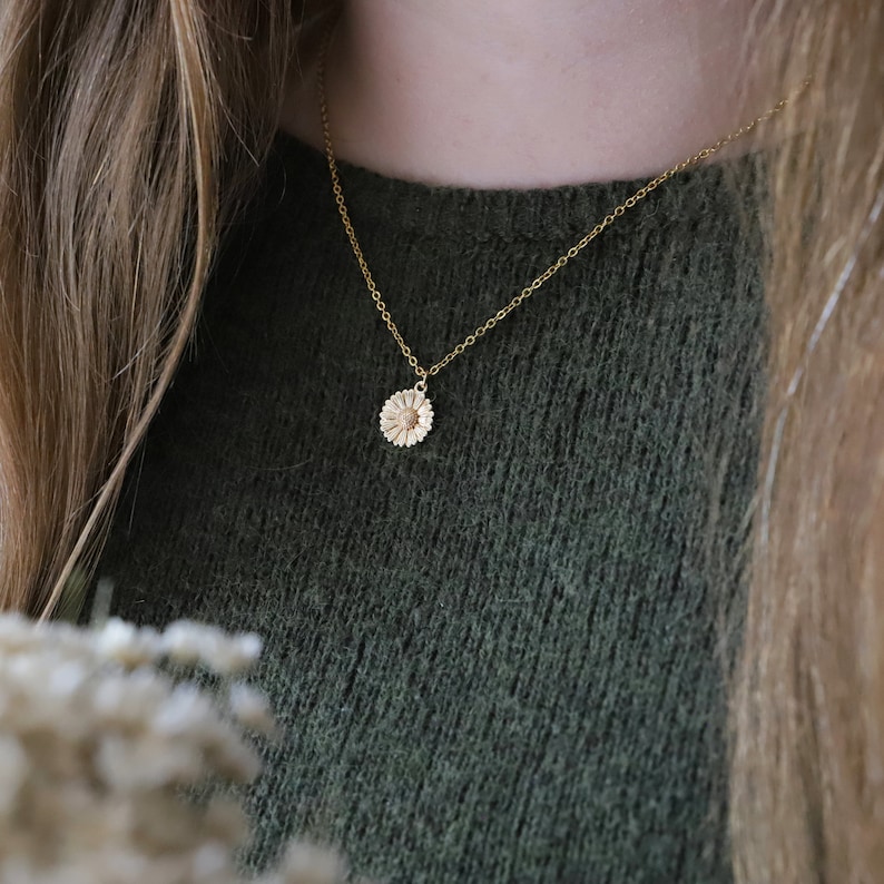 Puede incluir: Un collar de cadena dorada con un delicado colgante de margarita. El collar lo lleva una persona con el pelo largo y casta&ntilde;o y un jersey verde.