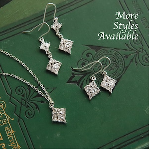 May include: A silver necklace and earrings set with a square pendant and matching earrings. The jewelry features clear cubic zirconia stones arranged in a floral pattern. The jewelry is displayed on a green book with the text "More Styles Available".