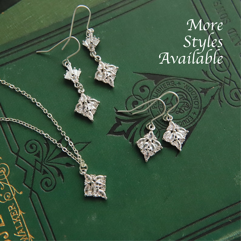 May include: A silver necklace and earrings set with a square pendant and matching earrings. The jewelry features clear cubic zirconia stones arranged in a floral pattern. The jewelry is displayed on a green book with the text "More Styles Available".