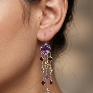 May include: Silver dangle earrings featuring a heart-shaped purple gemstone at the top. Below, a cascade of various colored gemstones, including red, yellow, and green, hangs from delicate chains. The earrings are shown against a blurred background.