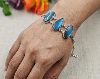 May include: A silver bracelet featuring three blue labradorite stones. The stones are oval and teardrop-shaped, set in silver bezels, and connected by a delicate chain. The bracelet is displayed on a wrist, with colorful flowers in the background.