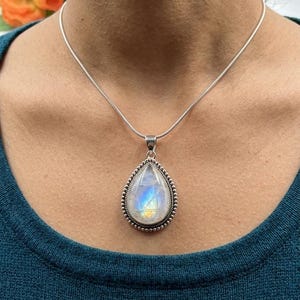 May include: A silver necklace featuring a pear-shaped pendant with a rainbow moonstone. The pendant is framed by a beaded silver border. The necklace is worn on a person wearing a teal-colored top.