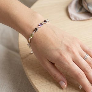 May include: A silver chain bracelet featuring a series of small, oval-shaped, multi-colored gemstones in shades of purple, green, yellow, and dark red. The bracelet is worn on the wrist, and a simple silver band ring is visible on the ring finger.