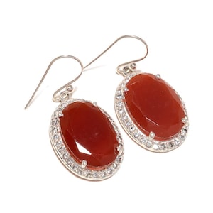 May include: A pair of oval-shaped earrings with a faceted, reddish-orange gemstone center. The gemstones are surrounded by a halo of small, clear crystals set in a silver-colored metal. The earrings have a hook closure.