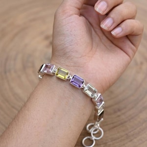 Puede incluir: Una pulsera de plata con piedras preciosas rectangulares en varios colores, incluyendo púrpura, amarillo y rosa. La pulsera se muestra en una muñeca sobre un fondo de madera. Las piedras están engarzadas en una banda de plata.