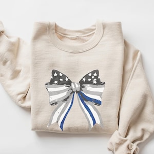 May include: A beige sweatshirt with a large bow printed on the front. The bow is made of an American flag pattern with a blue and white thin blue line ribbon.