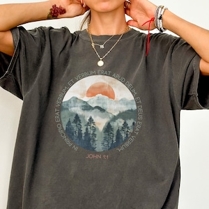 May include: A dark gray oversized t-shirt with a graphic print of a mountain landscape and a sunset. The text "IN PRINCIPIO ERAT VERBUM ET VERBUM ERAT APUD DEUM, ET DEUS ERAT VERBUM" and "JOHN 1:1" are printed on the graphic.