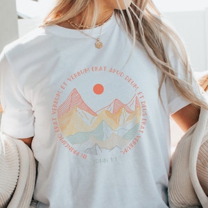 May include: White t-shirt with a graphic of colorful mountains and a sun. The text "In principio erat Verbum. John 1:1" is printed on the shirt.