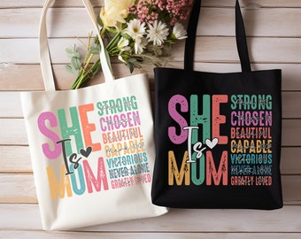 May include: Two tote bags with the text "She is Mom" in colorful letters. The text is layered with words like "strong", "chosen", "beautiful", "capable", "victorious", "never alone", and "greatly loved". One bag is white and the other is black.