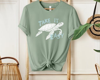 May include: Sage green t-shirt featuring a white sea turtle graphic and the text "Take it easy" in blue lettering.