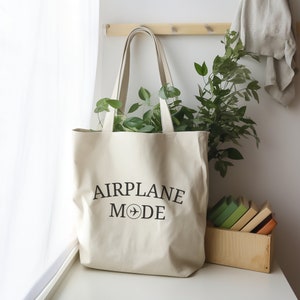 May include: A white canvas tote bag with black text that reads "AIRPLANE MODE" and a small airplane icon. The bag is sitting on a white surface with a potted plant and a wooden box of books behind it.