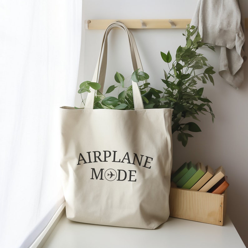 May include: A white canvas tote bag with black text that reads "AIRPLANE MODE" and a small airplane icon. The bag is sitting on a white surface with a potted plant and a wooden box of books behind it.