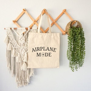 May include: A beige canvas tote bag with black text that reads "AIRPLANE MODE" and a small airplane icon. The bag is hanging on a wooden wall rack.