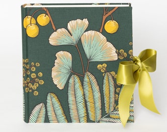 Puede incluir: Un álbum de fotos cubierto de tela verde con una cinta amarilla atada alrededor. El álbum presenta un patrón floral con hojas amarillas, verdes y blancas.