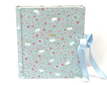 Puede incluir: Un álbum de fotos cuadrado con una cubierta azul claro con un estampado de conejos blancos, flores rosas y detalles verdes. El álbum está atado con una cinta azul claro. La palabra "Nancy" está impresa en la portada.