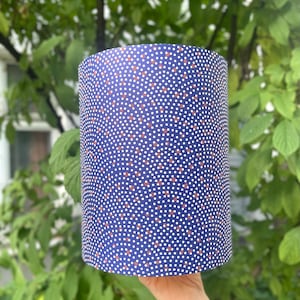 Könnte beinhalten: Ein zylindrischer Lampenschirm mit marineblauem Hintergrund und einem Muster aus kleinen weißen und braunen Punkten. Der Lampenschirm wird vor einem Hintergrund aus grünem Laub gehalten.