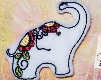 May include: White felt applique of an elephant with a floral design on its head. The elephant is outlined in black thread and has a red, yellow, and green floral design. The applique is made by Tatted Stitch Embroideries.