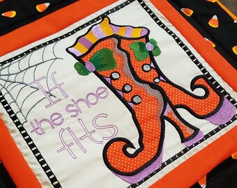 May include: A white fabric square with an orange and green witch's shoe embroidered on it. The text "If the shoe fits" is embroidered in purple. The square is framed with a black and white border and a red border.
