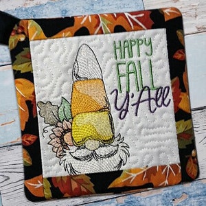 Puede incluir: Un agarrador de olla de tela negro y naranja con fondo blanco y un diseño de gnomo de maíz dulce. El texto "Happy Fall Y'all" está bordado en verde y morado.