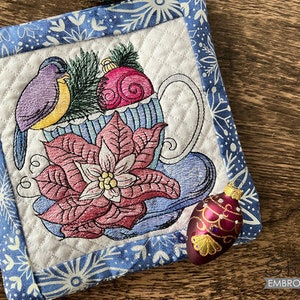 May include: A blue and white quilted teacup with a poinsettia and a blue bird with a red ornament. The teacup is on a white background with a blue border.