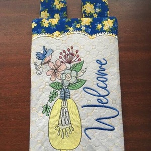 Può includere: Un appendiporta in tessuto trapuntato bianco con un motivo floreale blu nella parte superiore. Il centro dell'appendiporta presenta un disegno ricamato di una lampadina con fiori e un uccello. La parola "Benvenuto" è ricamata in lettere corsive blu.