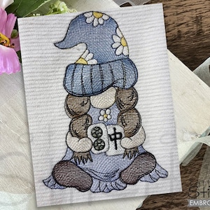 May include: Embroidered gnome design in blue, brown, and white. The gnome wears a blue hat with white daisies and a blue outfit. The design is on a white background with the text "Tattered Stitch Embroideries".