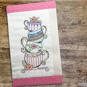 May include: A stack of four teacups with floral designs embroidered on a pink and white fabric with a polka dot border. The fabric has the text "Embroideries" on the bottom right corner.