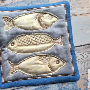 Può includere: Un sottopentola in tessuto blu e marrone con tre pesci ricamati in beige e marrone su uno sfondo blu chiaro.