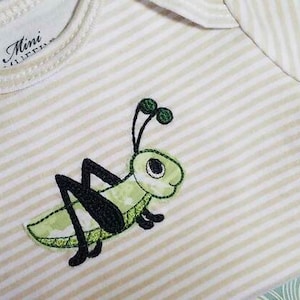 May include: A green and black embroidered grasshopper design on a white and beige striped fabric. The design is for a baby onesie.